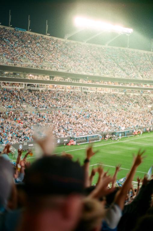 A stadium full of people is visible from behind the heads and raised hands of a crowd in the foreground.