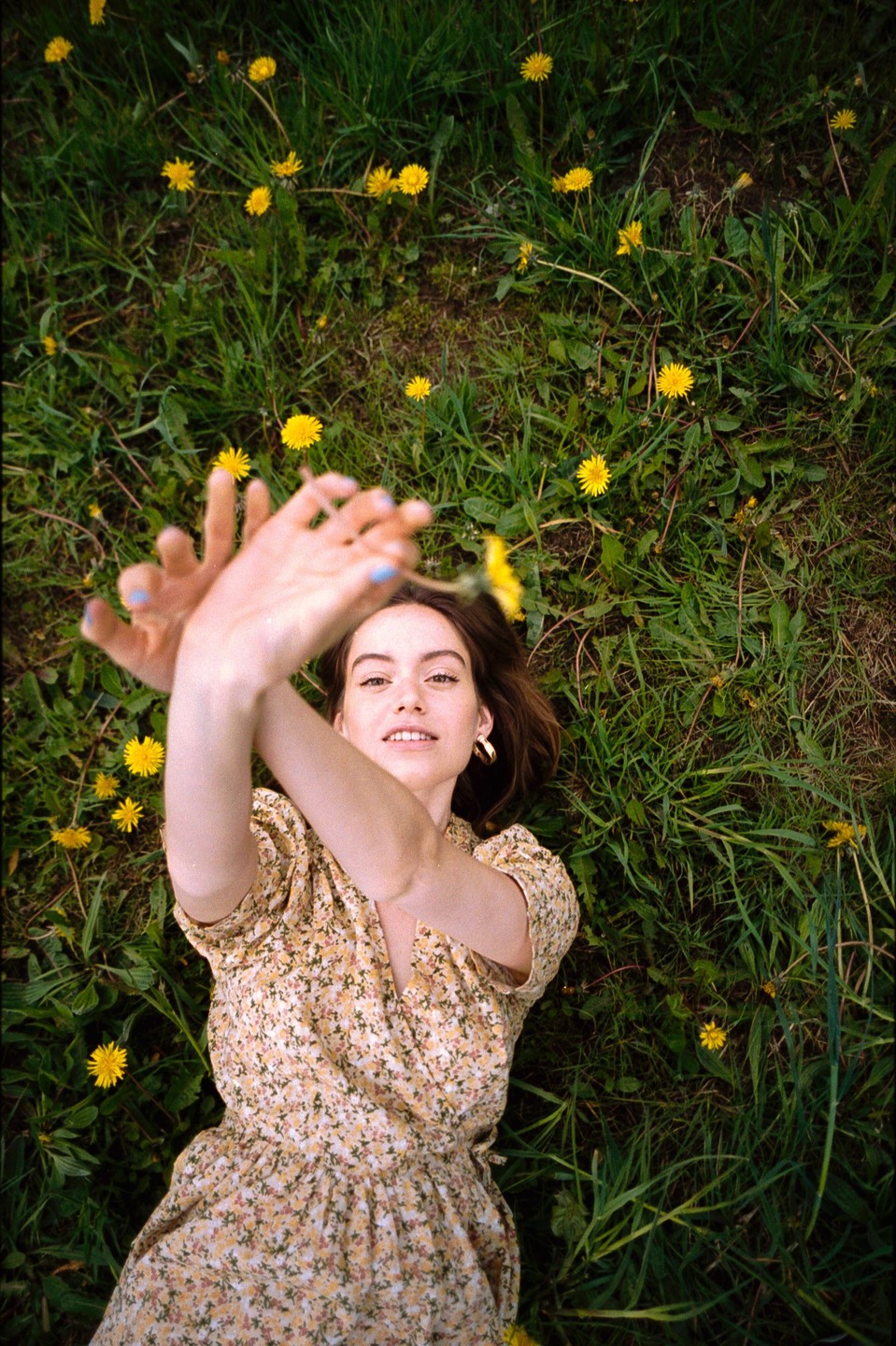 A woman in a floral dress lies in a field of dandelions, looking up at the camera with her arms raised.