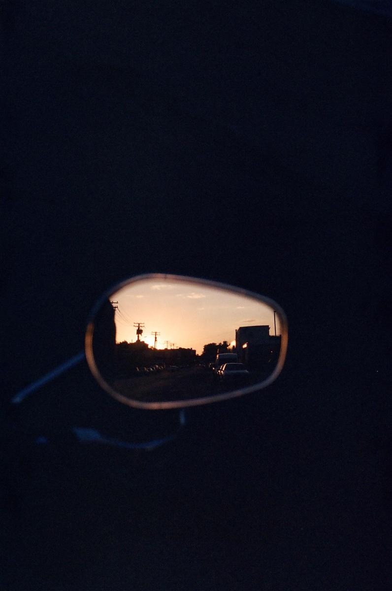 A sunset is reflected in a vehicle's side mirror, showing a street with cars and utility poles.