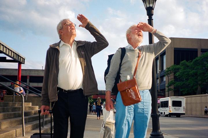 Two men shield their eyes from the sun while standing on a street next to a lamppost.