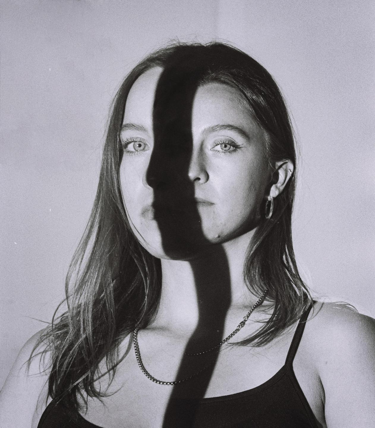 A woman with long hair and a necklace is illuminated by a strong shadow across her face.