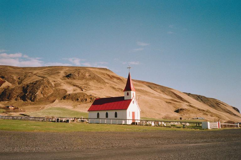 A white church with a red roof stands in a green field with a brown hill behind it under a clear blue sky.