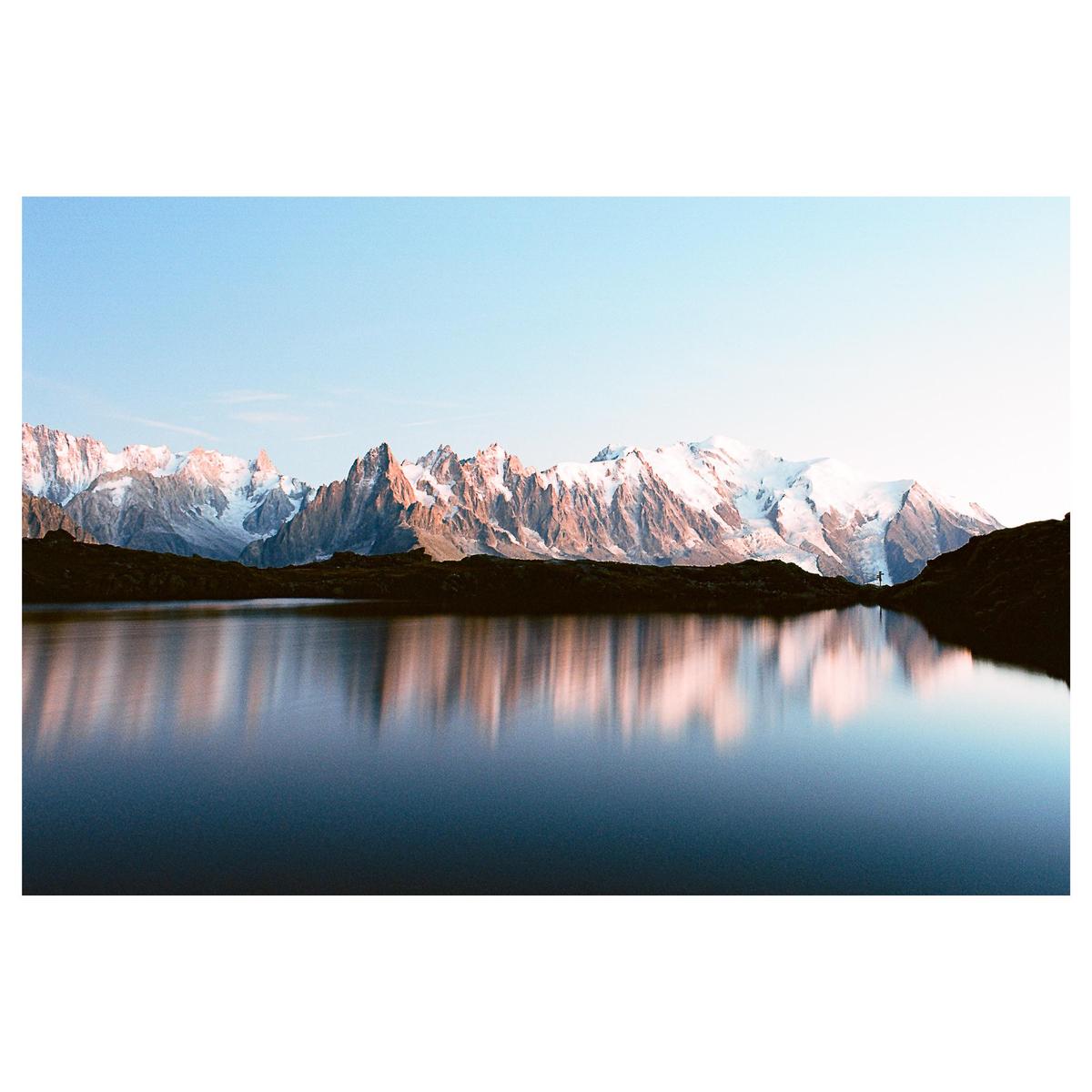 A mountain range with snow-capped peaks is reflected in a calm lake under a clear sky.
