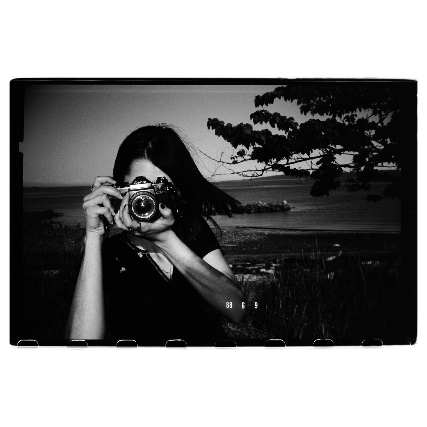 A woman holds a camera to her face, with a body of water and trees in the background.
