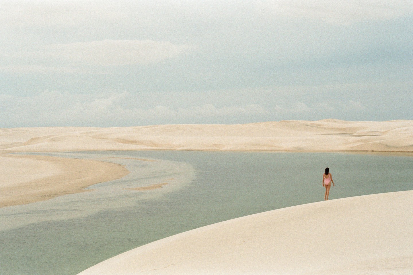 A woman in a pink swimsuit walks towards a lagoon surrounded by sand dunes under a cloudy sky.
