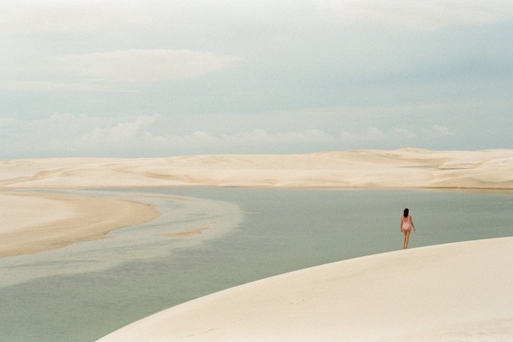A woman in a pink swimsuit walks towards a lagoon surrounded by sand dunes under a cloudy sky.