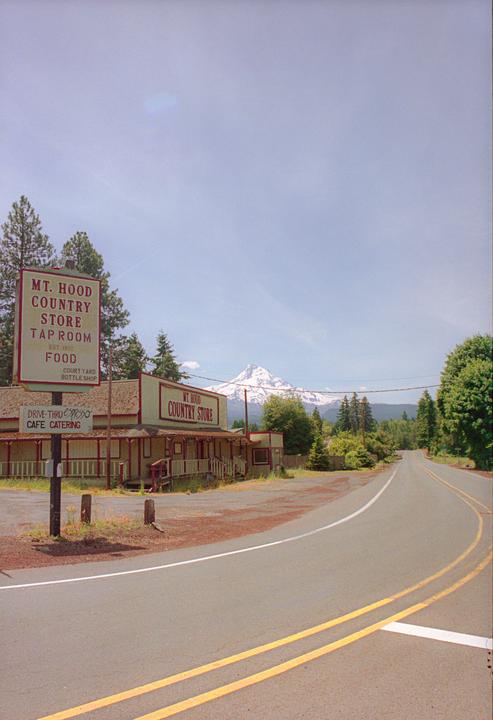 A country store with a large sign stands beside a road, with a snow-capped mountain visible in the distance.
