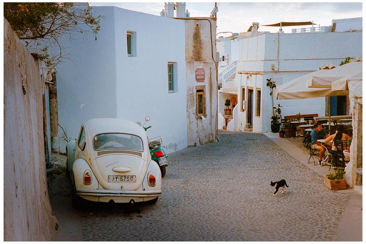 A white beetle car is parked on a cobblestone street next to a white building, with a black cat walking in the foreground and a couple sitting at a cafe table in the background.