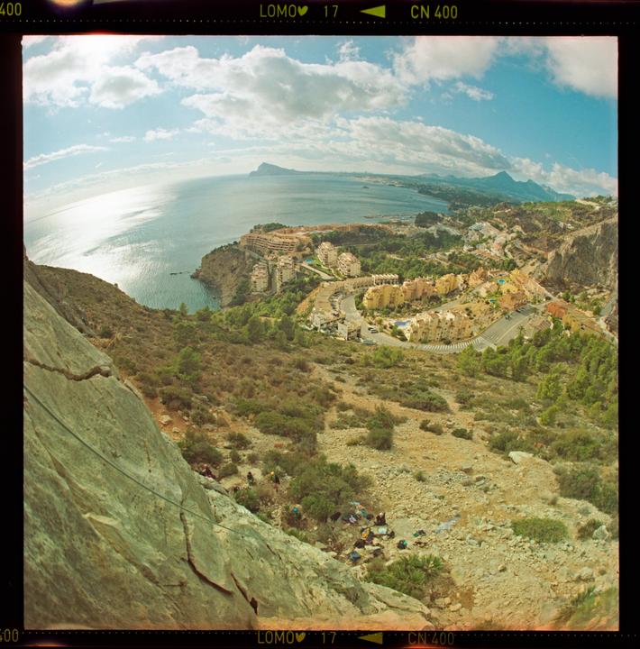A wide-angle landscape shows a rocky mountain slope overlooking a coastal town with buildings nestled along the sea, under a bright sky with clouds.