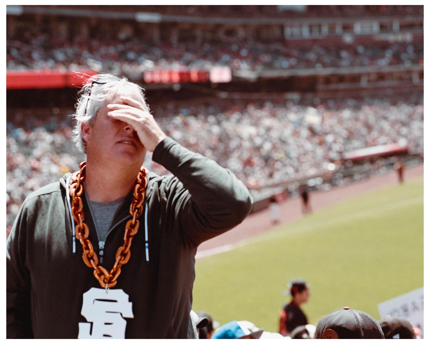 A man wearing a large chain necklace covers his face with his hand at a baseball game.