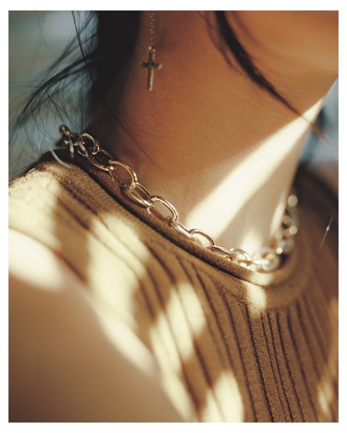 A woman wears a gold chain necklace and a cross earring, with sunlight casting shadows on her ribbed top.