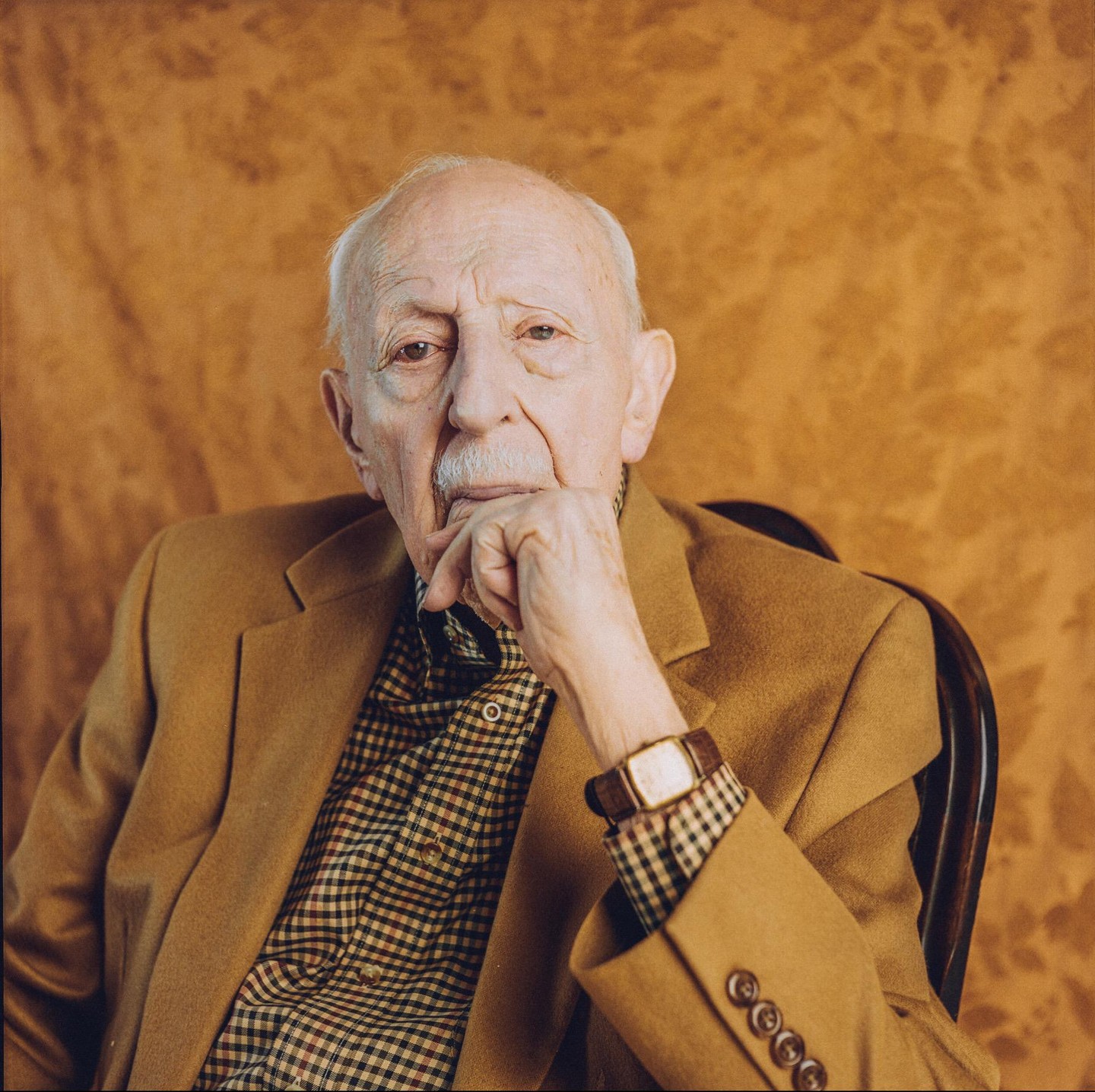 A man with a mustache sits in a chair, resting his chin on his hand.
