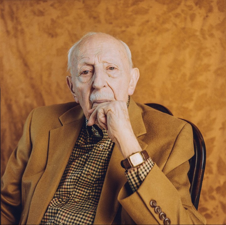 A man with a mustache sits in a chair, resting his chin on his hand.