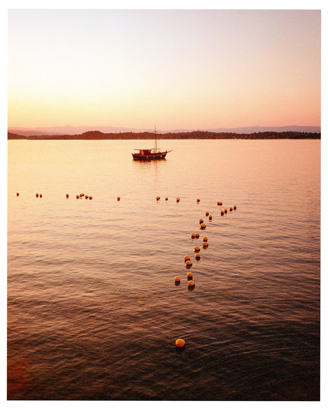 A boat floats on the water with a line of buoys leading towards it at sunset.