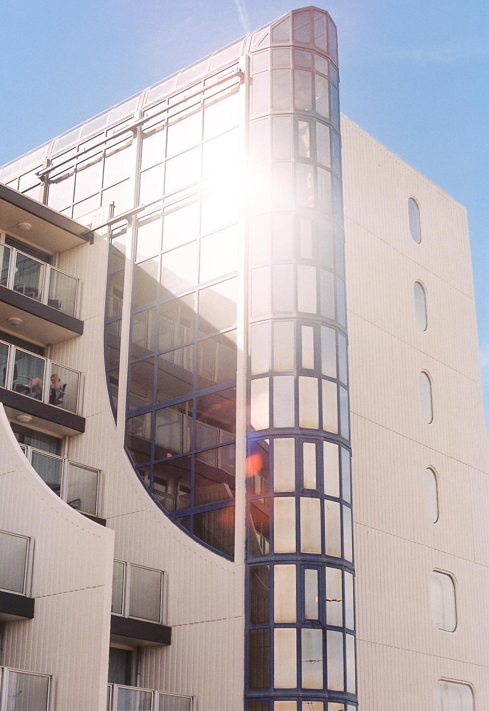 The sun reflects brightly off the glass facade of a modern building with balconies and a curved glass tower.