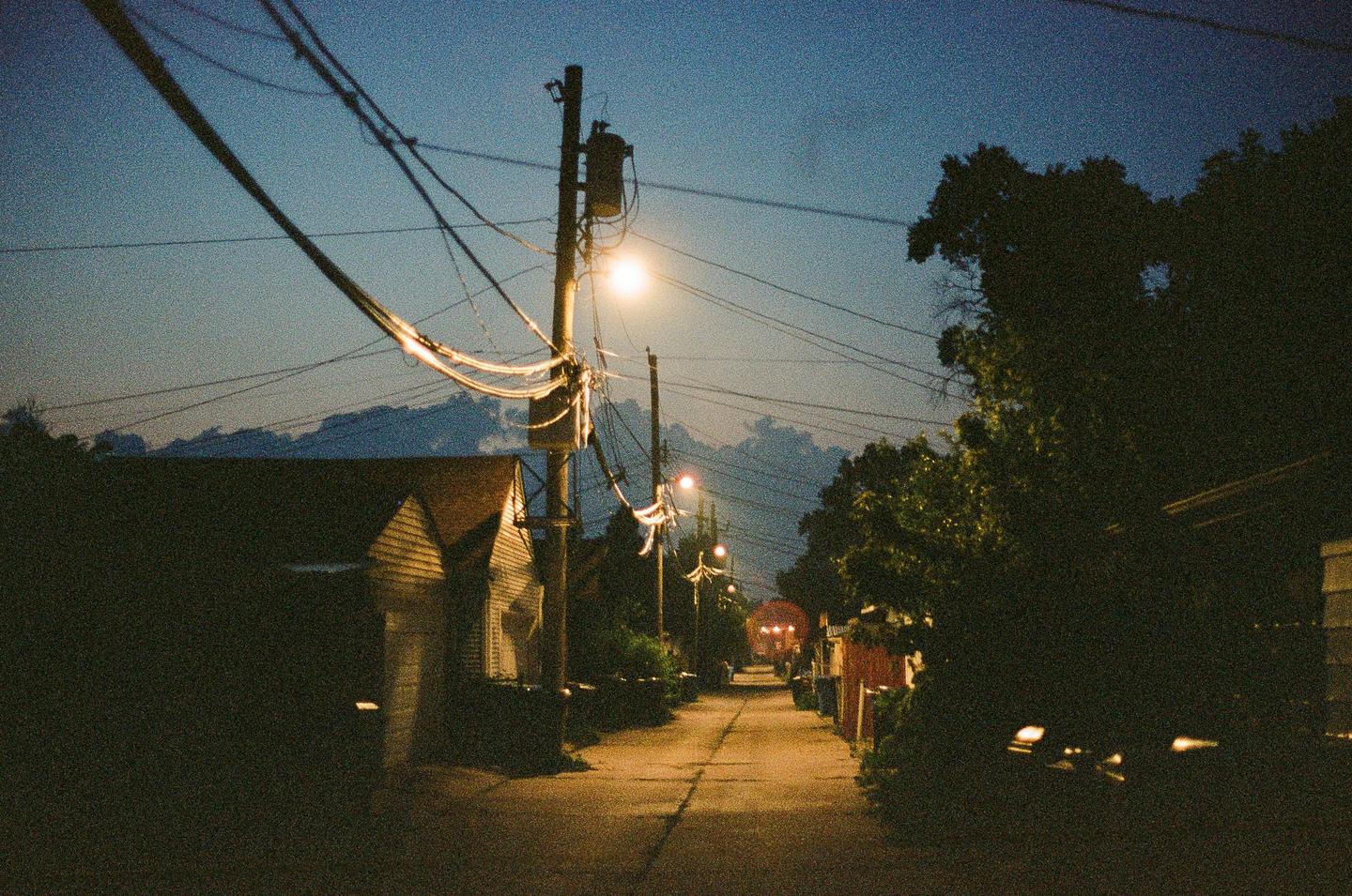 A narrow alleyway is illuminated by streetlights at night, with houses and trees lining its sides.