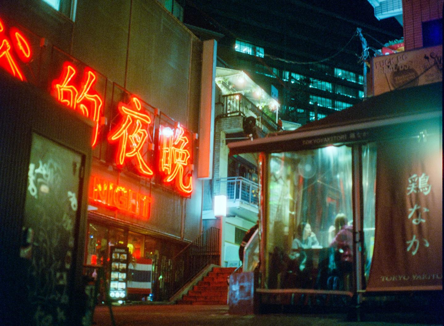 A street at night is illuminated by red neon signs and the lights of a building.