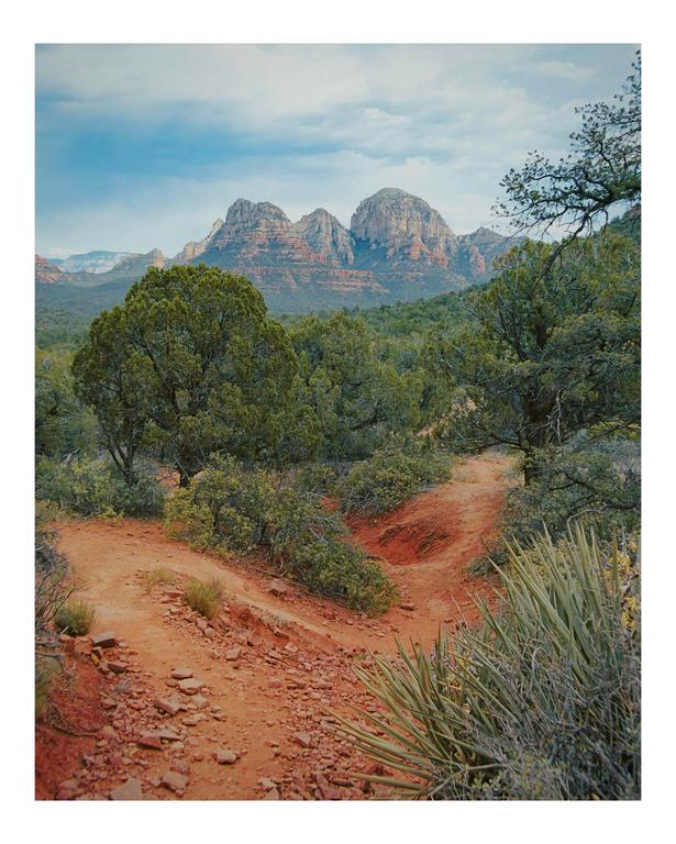 A dirt path winds through a desert landscape with green trees and shrubs, leading towards large red rock mountains under a cloudy sky.