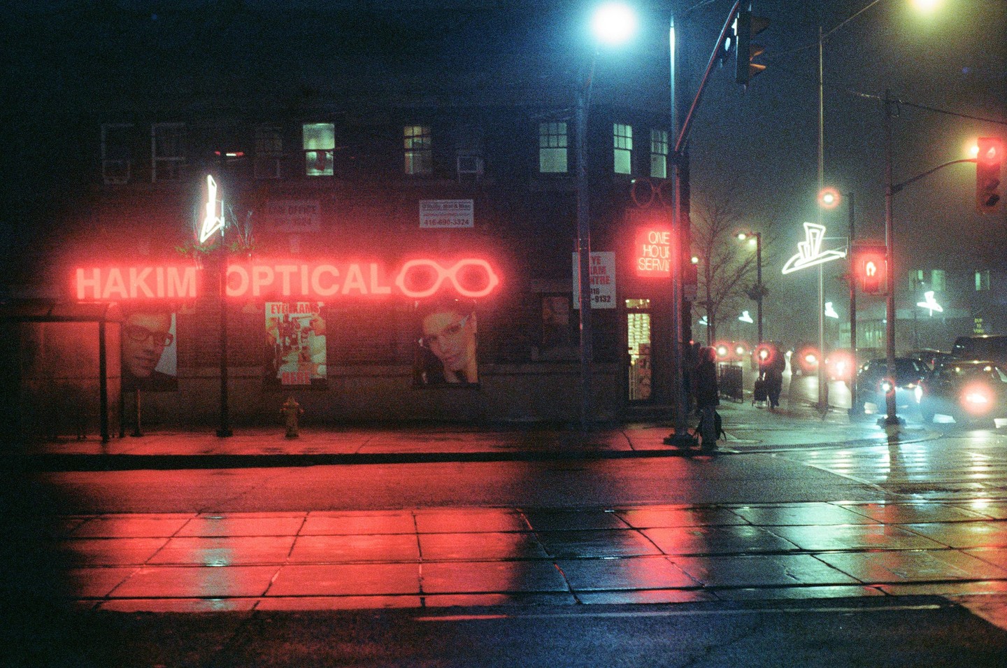 A street corner at night is illuminated by the red neon sign of an optical store and the headlights of cars.
