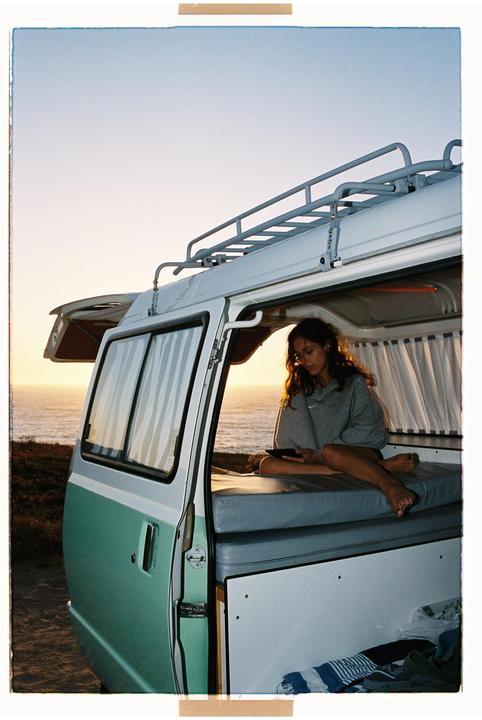 A woman sits in the back of a camper van looking at a tablet with the ocean visible in the background at sunset.