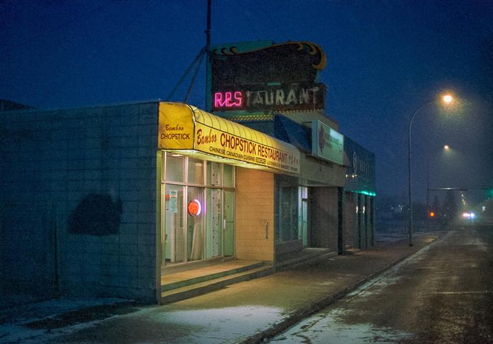 A restaurant with a yellow awning and a neon sign is visible on a street at night.