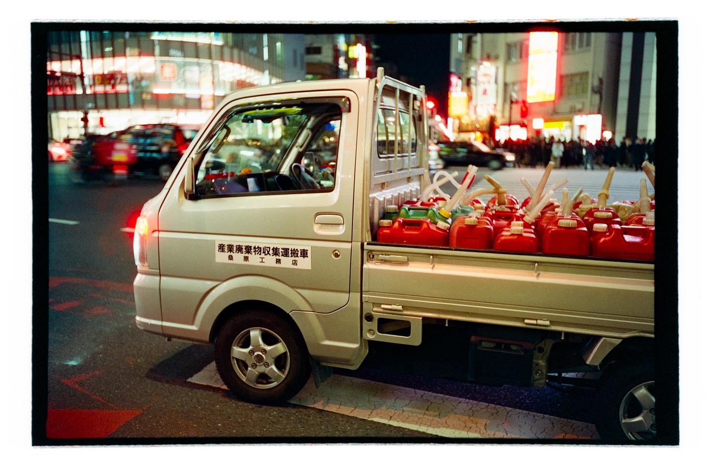 A small truck with many red and green gas cans in its bed is parked on a city street at night.