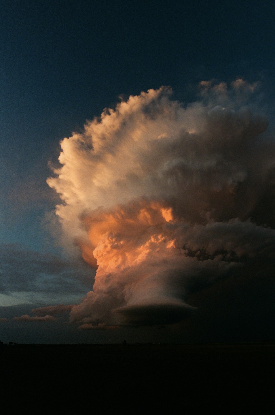 A large storm cloud with orange and white highlights dominates the sky above a dark landscape at sunset.