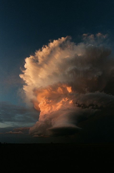 A large storm cloud with orange and white highlights dominates the sky above a dark landscape at sunset.