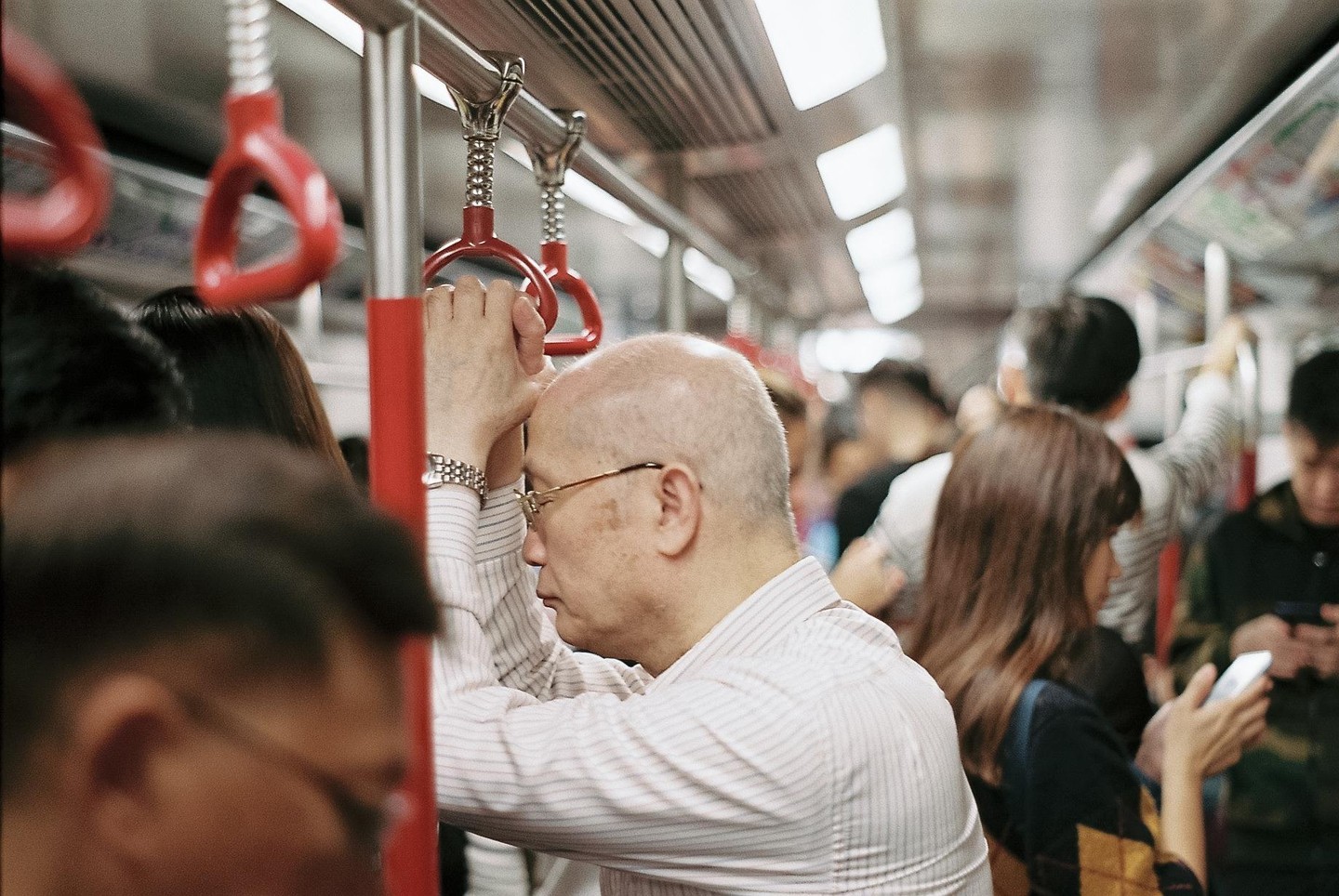A man with glasses leans against a pole, holding a red strap in a crowded subway car.