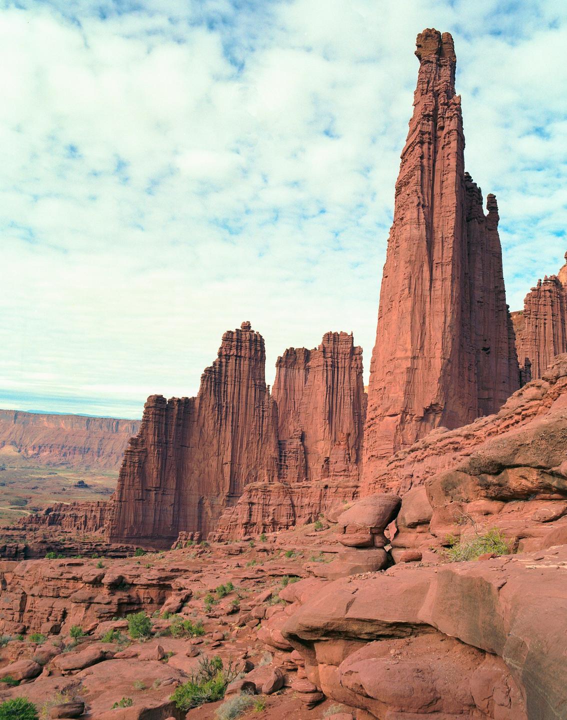 A tall red rock formation stands prominently against a cloudy sky, with other rock formations and a canyon visible in the background.