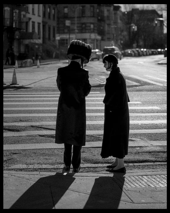 A man and a woman stand on a street corner with long shadows stretching across the sidewalk.