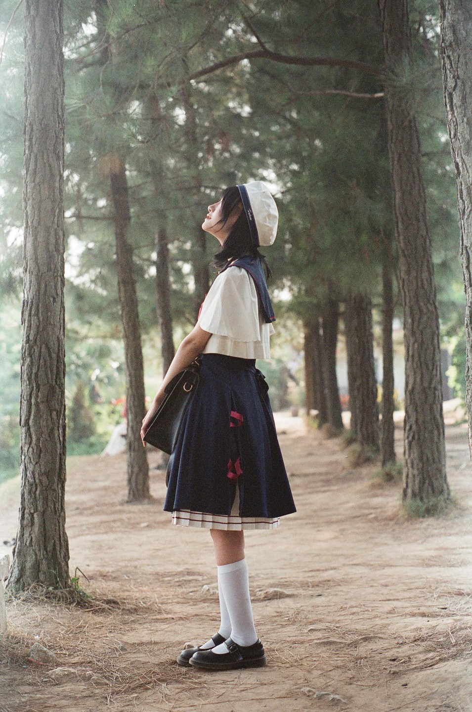 A girl in a school uniform stands in a forest looking up at the trees.