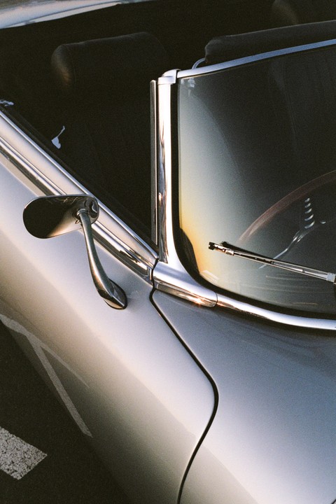 A close-up of a silver vintage car shows its side mirror, windshield, and chrome trim.