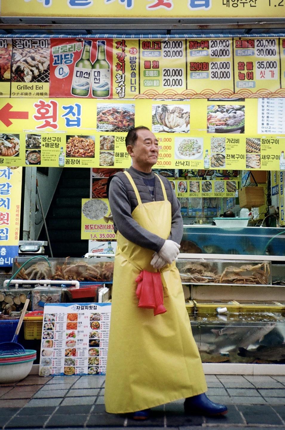 A man wearing a yellow apron and white gloves stands in front of a seafood stall with tanks of live seafood and colorful signs.