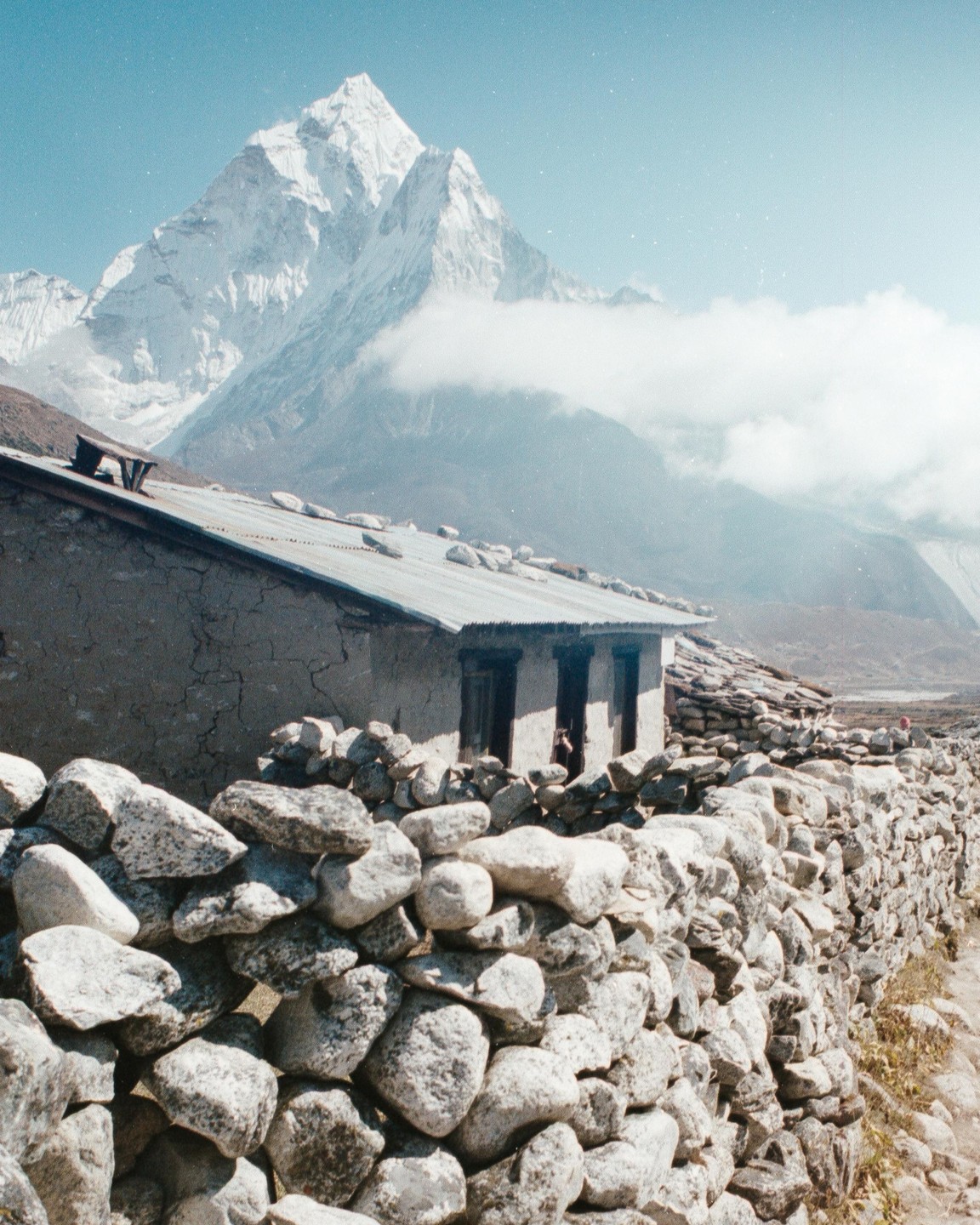 A stone wall and a building with a corrugated metal roof sit in the foreground of a snow-capped mountain partially obscured by clouds.