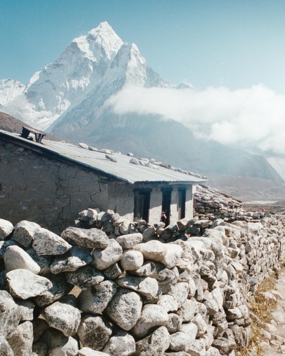 A stone wall and a building with a corrugated metal roof sit in the foreground of a snow-capped mountain partially obscured by clouds.