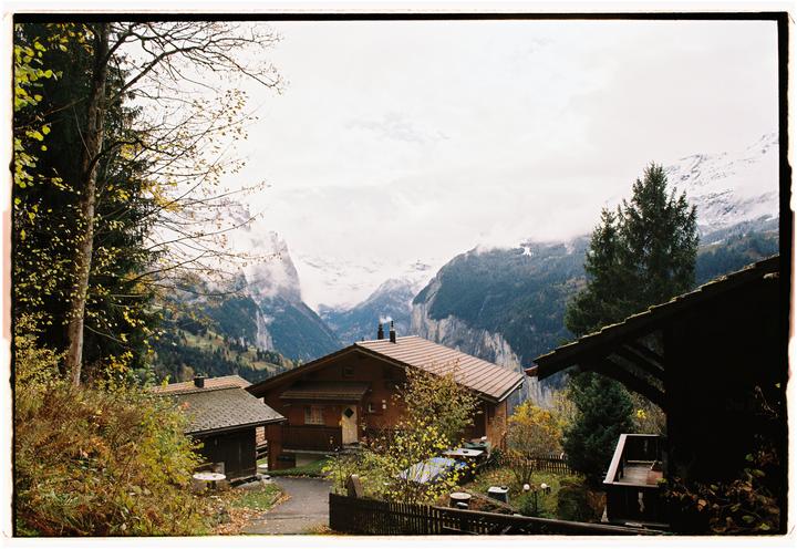 A wooden house with a brown roof sits in a valley surrounded by mountains and trees with autumn foliage.