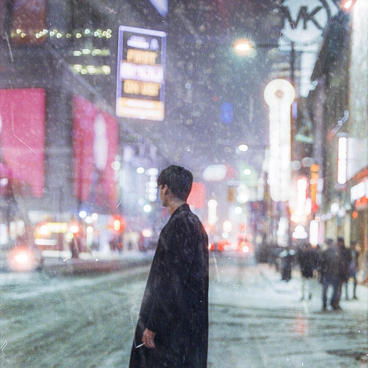 A man in a long coat stands on a snowy city street at night, looking at bright neon signs and distant traffic.