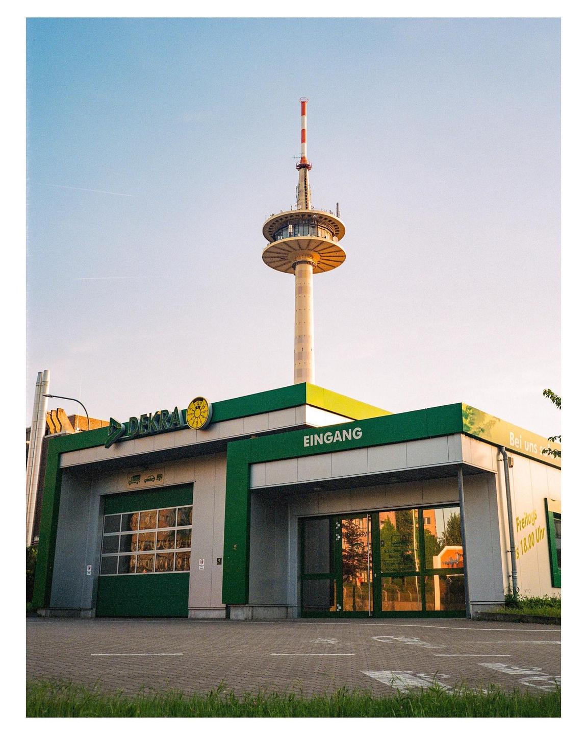 A tall communication tower stands behind a modern building with green accents and large windows.