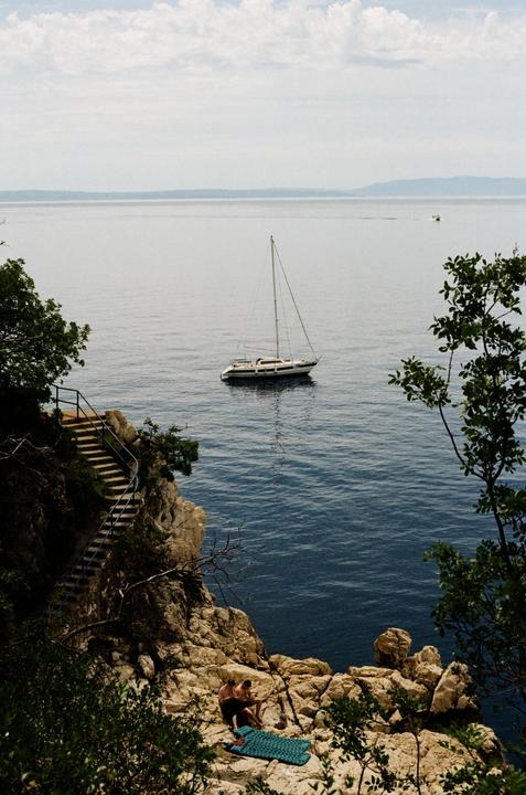A sailboat floats on the water near a rocky cliff with stairs leading down to the sea, where two people are sitting.
