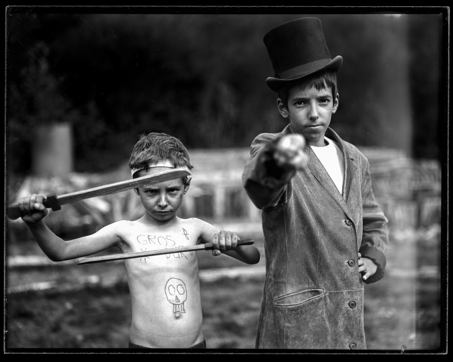Two boys pose for a portrait, one wearing a top hat and pointing a stick, the other holding two sticks and having a skull drawn on his stomach.