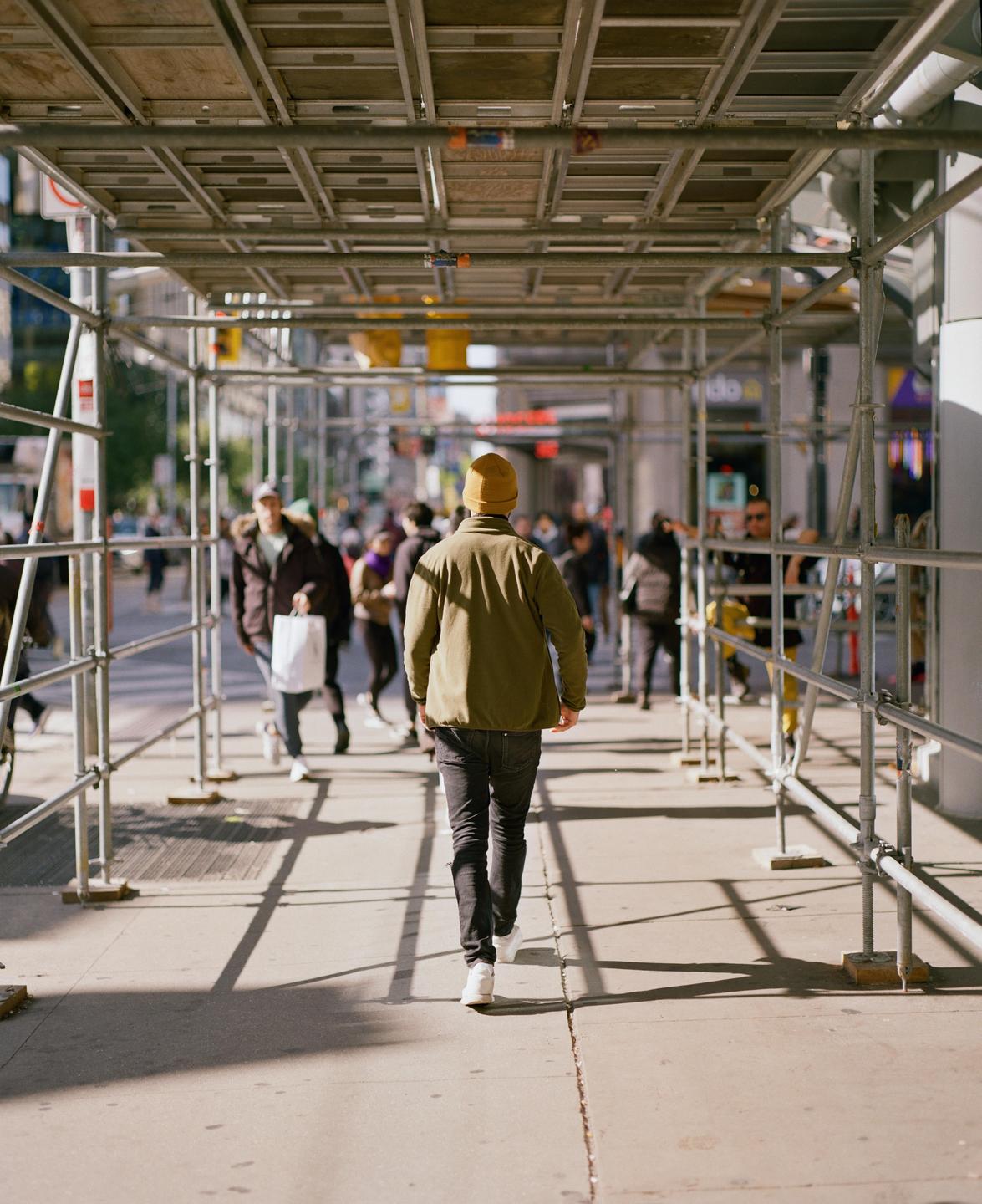 A man in a green jacket and yellow beanie walks away from the viewer under scaffolding on a city street.