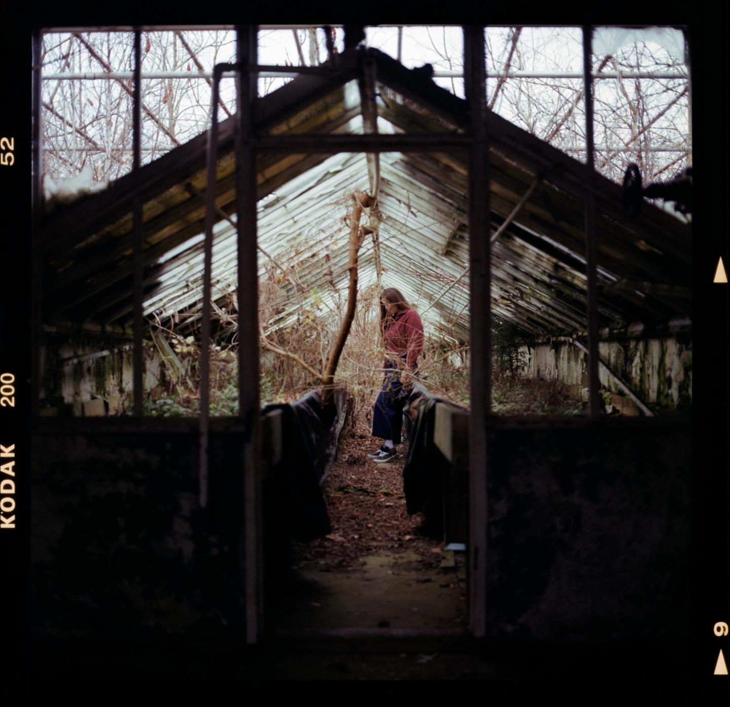 A woman stands inside a dilapidated greenhouse with broken glass panels and overgrown plants.