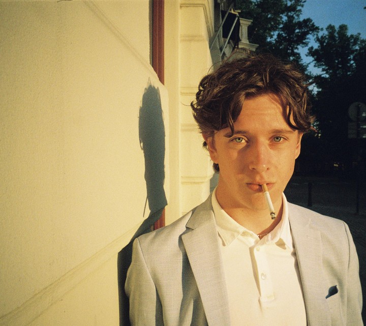 A man with a cigarette in his mouth stands next to a wall with a shadow.