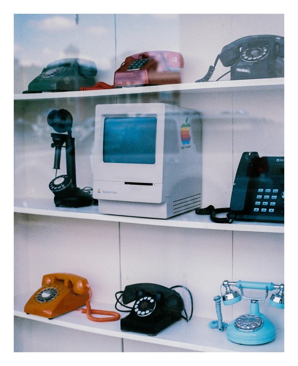 A collection of vintage telephones and a Macintosh Classic computer are displayed on white shelves.