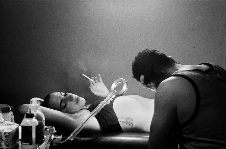 A woman lies on a table smoking a cigarette while a man tattoos her side.