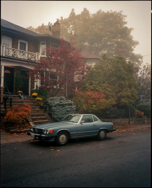 A blue car is parked on a street in front of a house with autumn foliage.