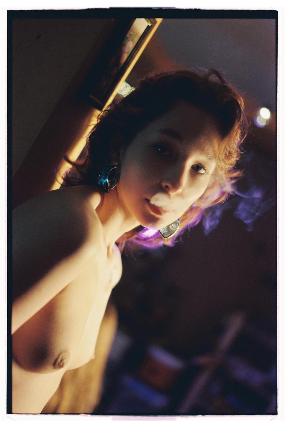 A woman with bare shoulders and large earrings looks at the camera while exhaling smoke, with purple light illuminating her hair.