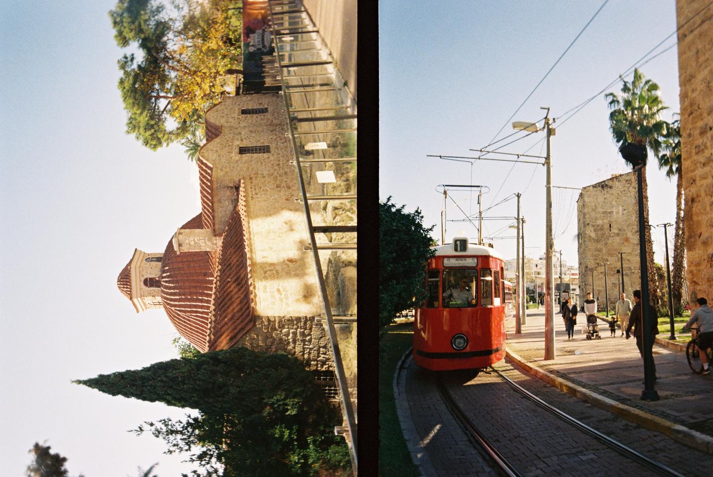 A red tram is on a track next to a stone building with a domed roof and a street with pedestrians.