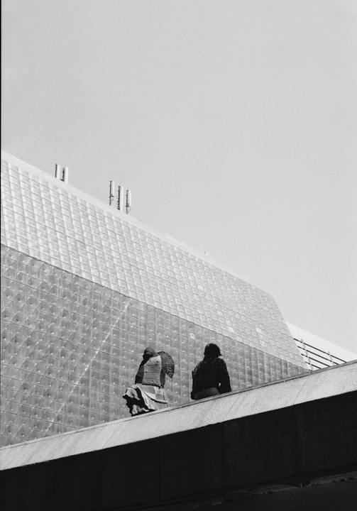 Two figures sit on a ledge in front of a glass block building.
